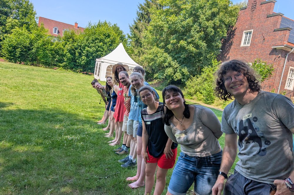 Ca. 10 Personen stehen in einer Reihe auf einer Wiese, leicht nach vorne geneigt, und lächeln in die Kamera